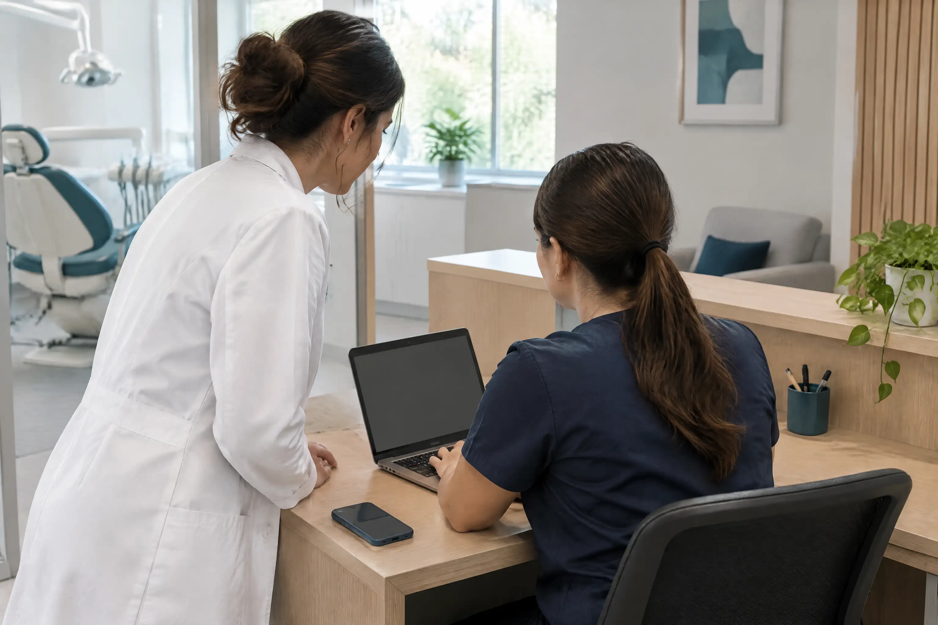 Dentista de jaleco e secretária olhando juntas o notebook na recepção do consultório.